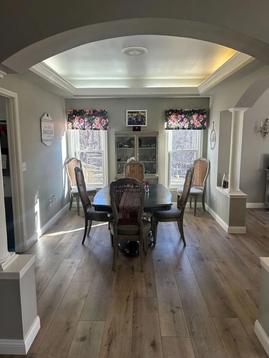 Dining room with a set table, wooden chairs, and bright windows with floral valances