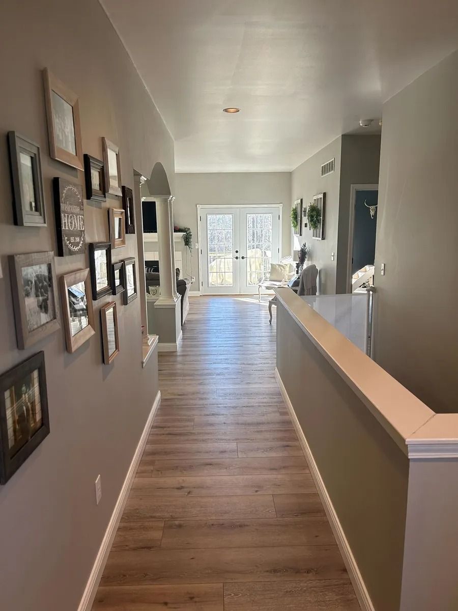 Narrow hallway with framed photos on the left and wood floors leading to a bright room with French doors.