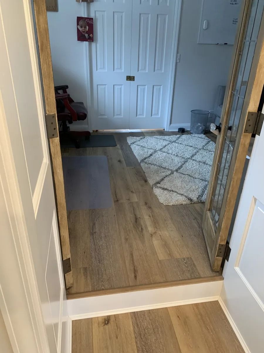 View through an open doorway into a hallway with wood flooring and a white door, partly covered by a rug.