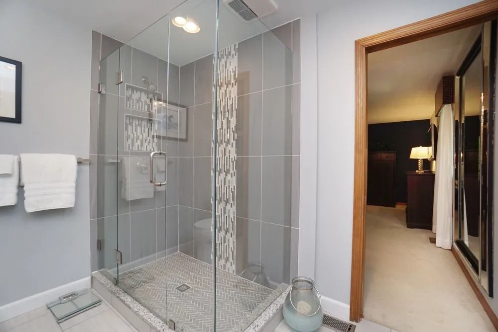 Glass walk-in shower with tiled floor and walls, next to a bathroom doorway and mirror.