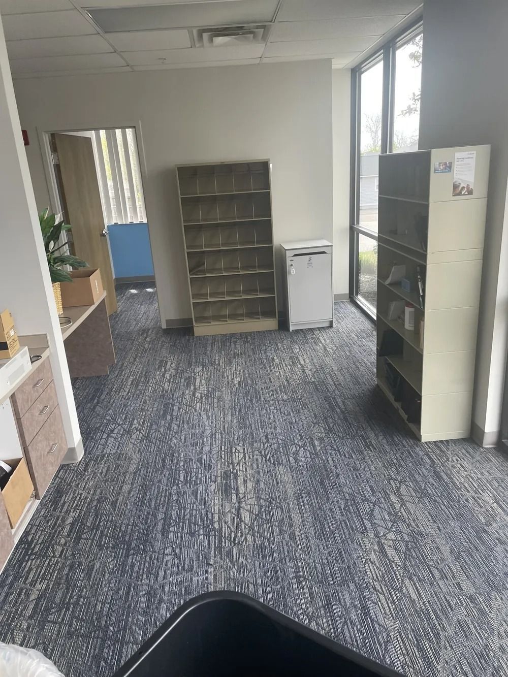 Empty office room with gray carpet, shelving units, and large windows along the right wall