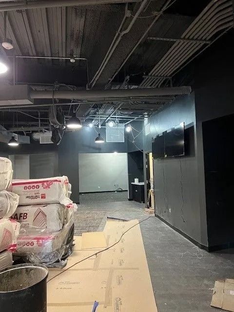 Industrial hallway with stacked wrapped materials, cardboard on the floor, and black walls under exposed ceiling lights