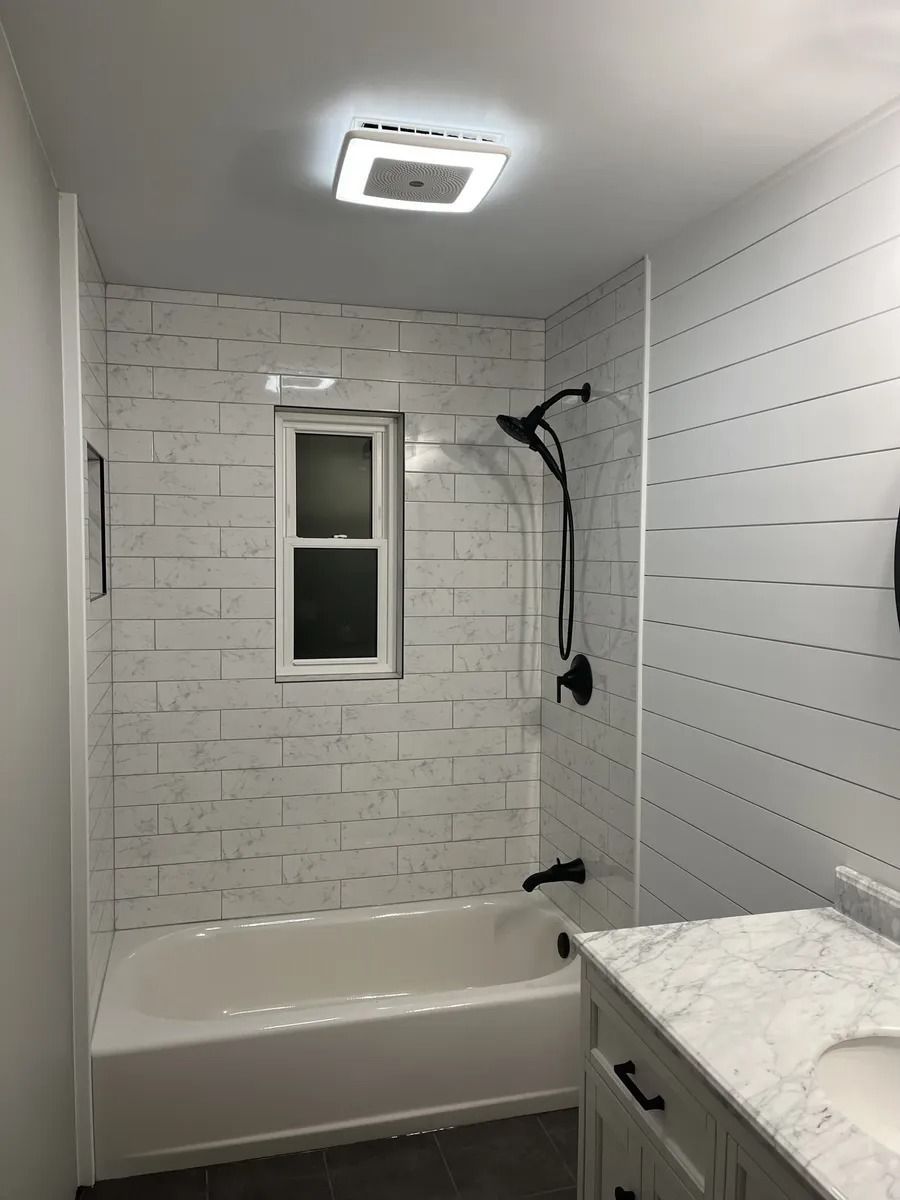 Modern white-tiled bathroom with a bathtub, window, showerhead, and marble vanity countertop