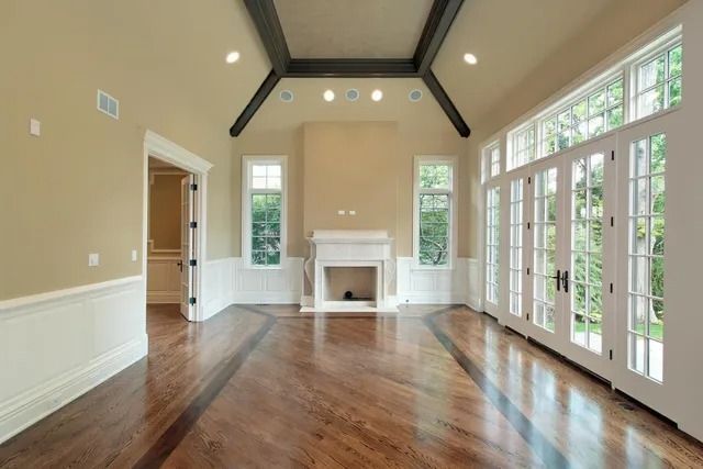 Empty living room with hardwood floors, white fireplace, tall windows, and vaulted ceiling with exposed beams
