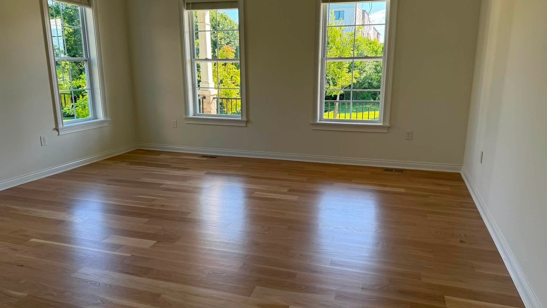 Empty sunlit room with hardwood floors and three tall windows overlooking greenery