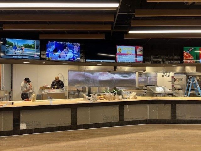 Empty cafeteria counter with two workers behind a glass serving line and TV screens overhead