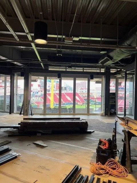 Empty interior with construction materials, exposed ceiling, and large windows overlooking a sports field.