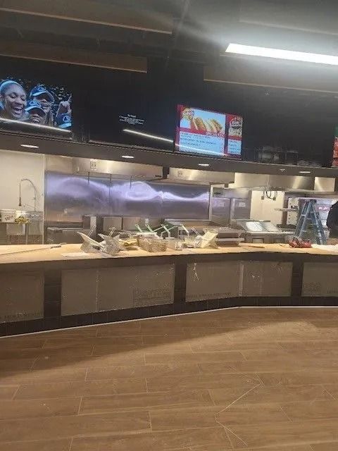 Empty restaurant buffet counter with stainless steel pans and overhead menu screens, in a dim dining area