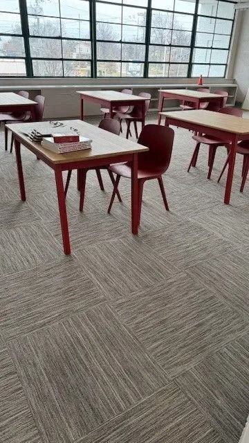 Empty classroom with red tables and chairs beside large windows, carpeted floor.