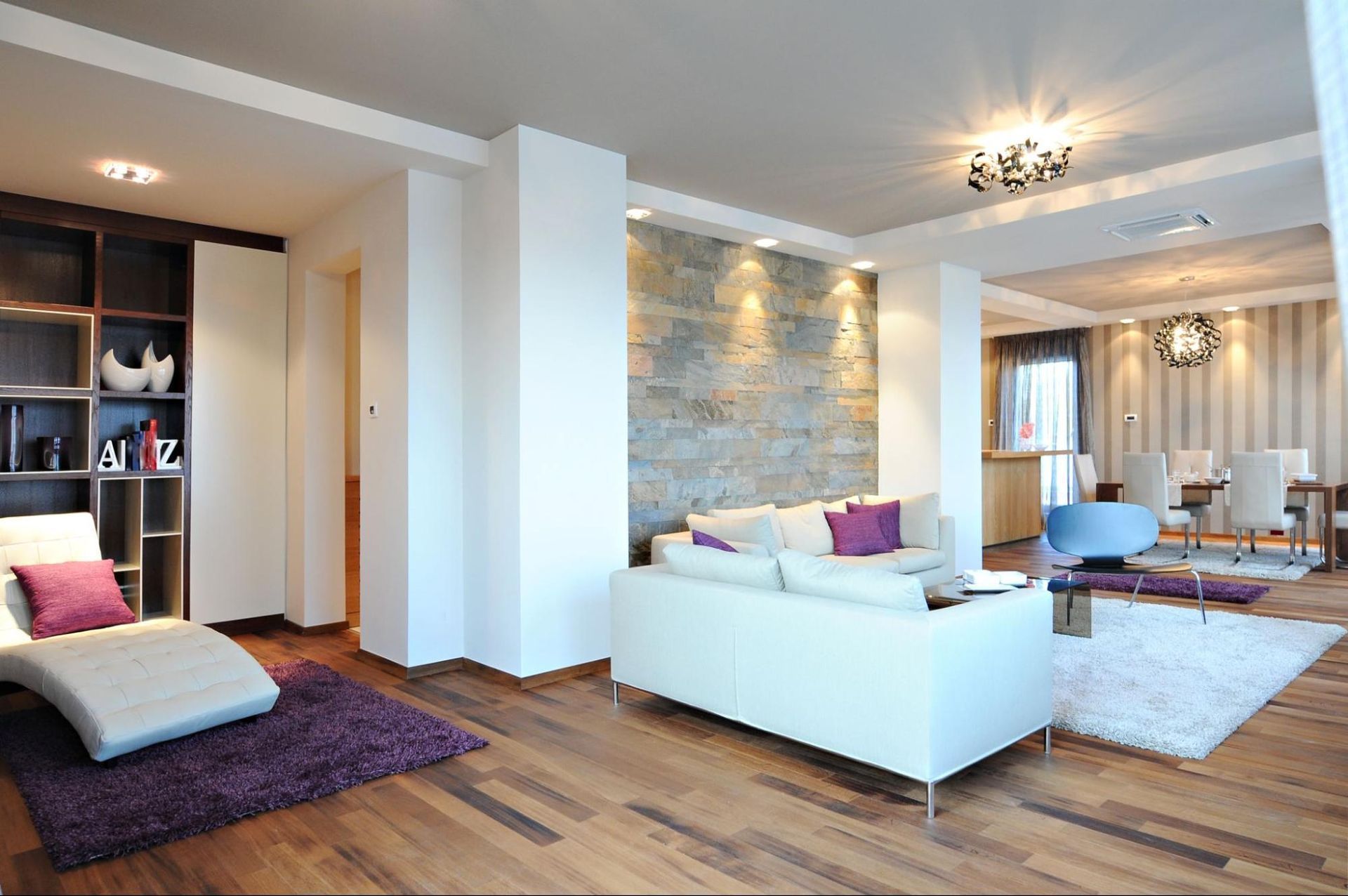 Modern open-plan living room with white sofas, purple accents, wooden floors, and stone feature wall