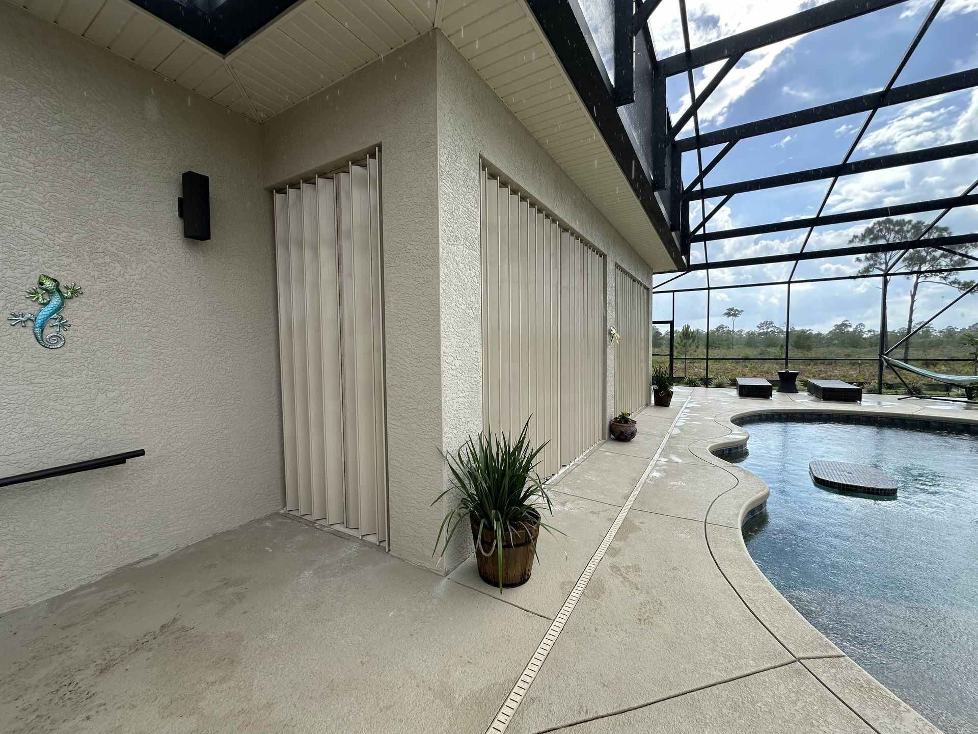 Screened pool patio beside a house, with sliding doors, potted plants, and blue water under a covered lanai.