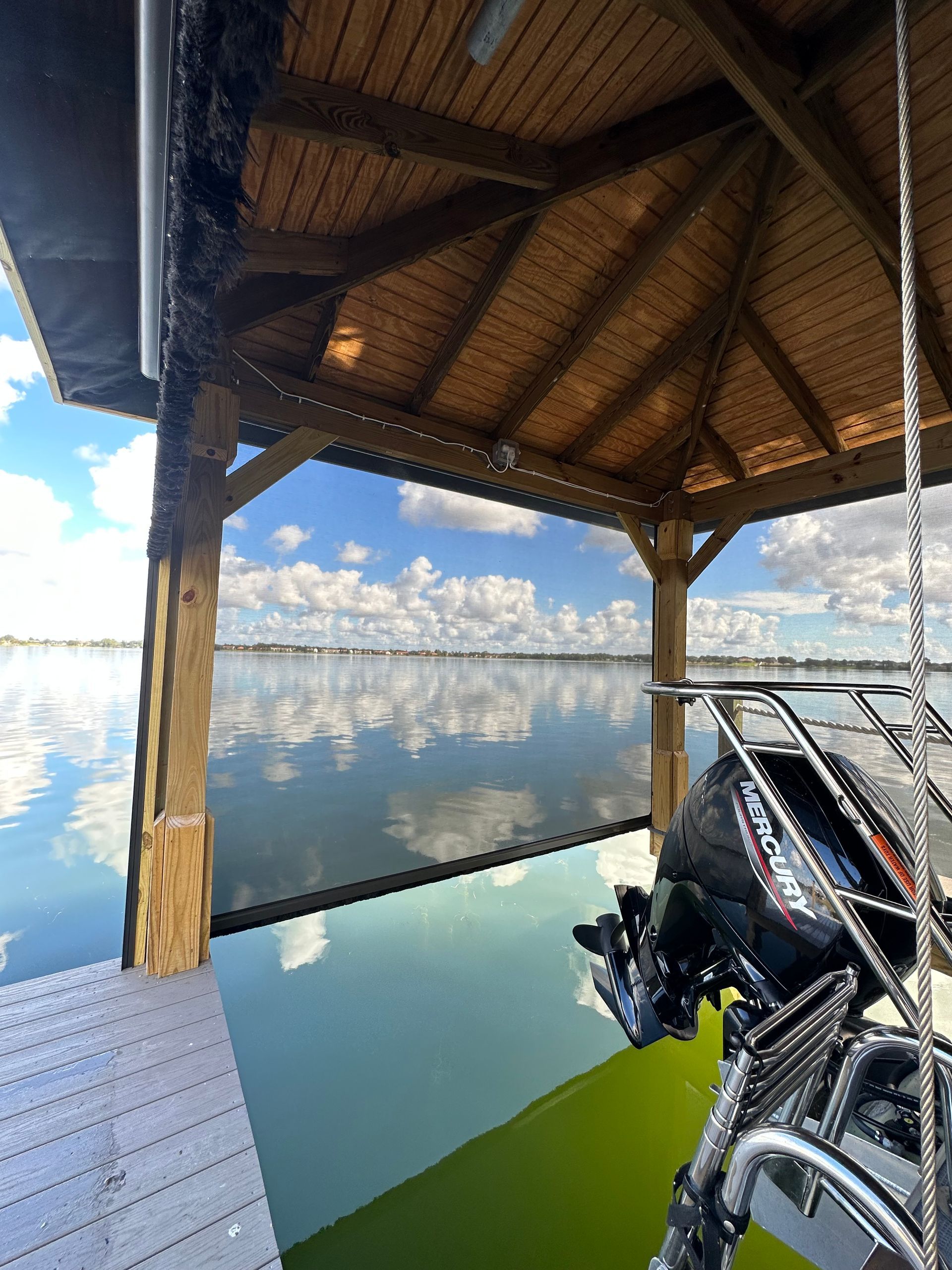 Wooden lakeside dock roof over calm water, with fishing gear and a reflection of clouds and sky.
