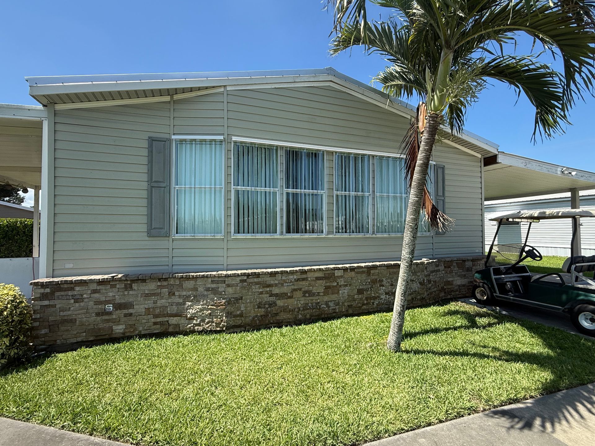 Single-story house with light siding, large front window, stone skirting, and a palm tree in the yard.