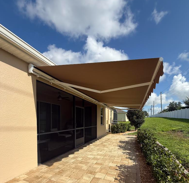 Patio with tan awning beside a beige house, paved walkway, and landscaped yard under a blue sky