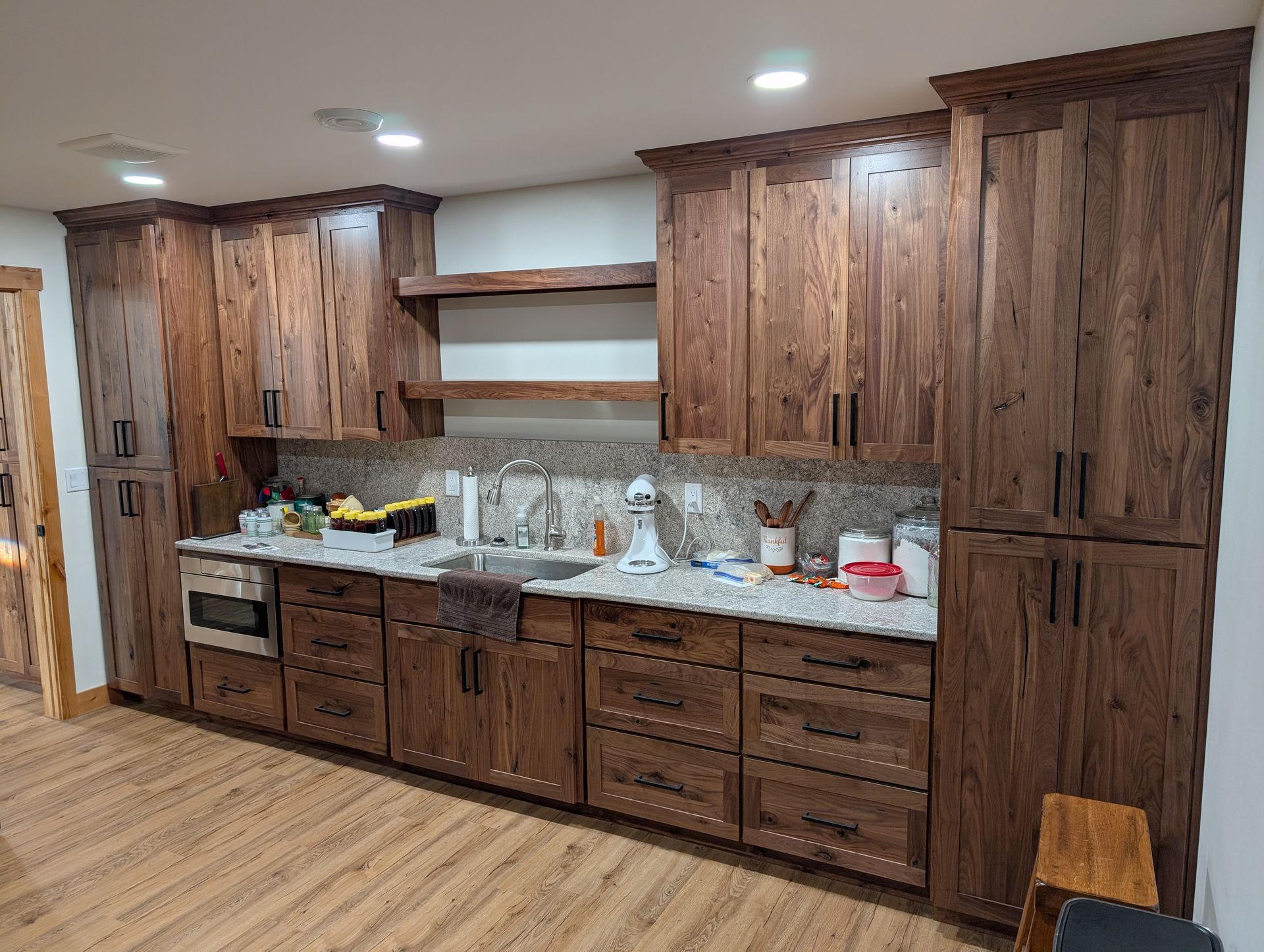 A kitchen featuring dark wood cabinets, light stone countertops, open shelving, and a stainless steel oven and sink.