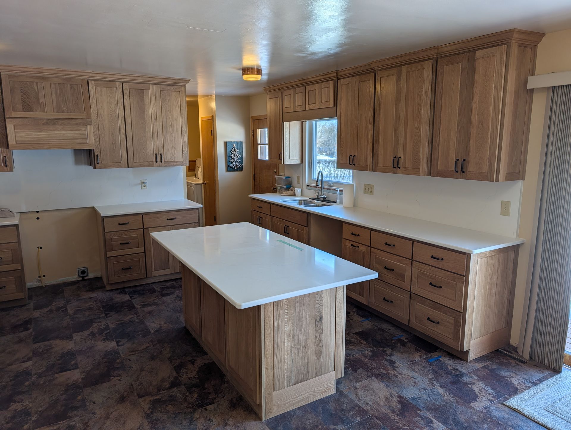 A renovated kitchen with light oak cabinets, white countertops, an island, and dark patterned flooring. 