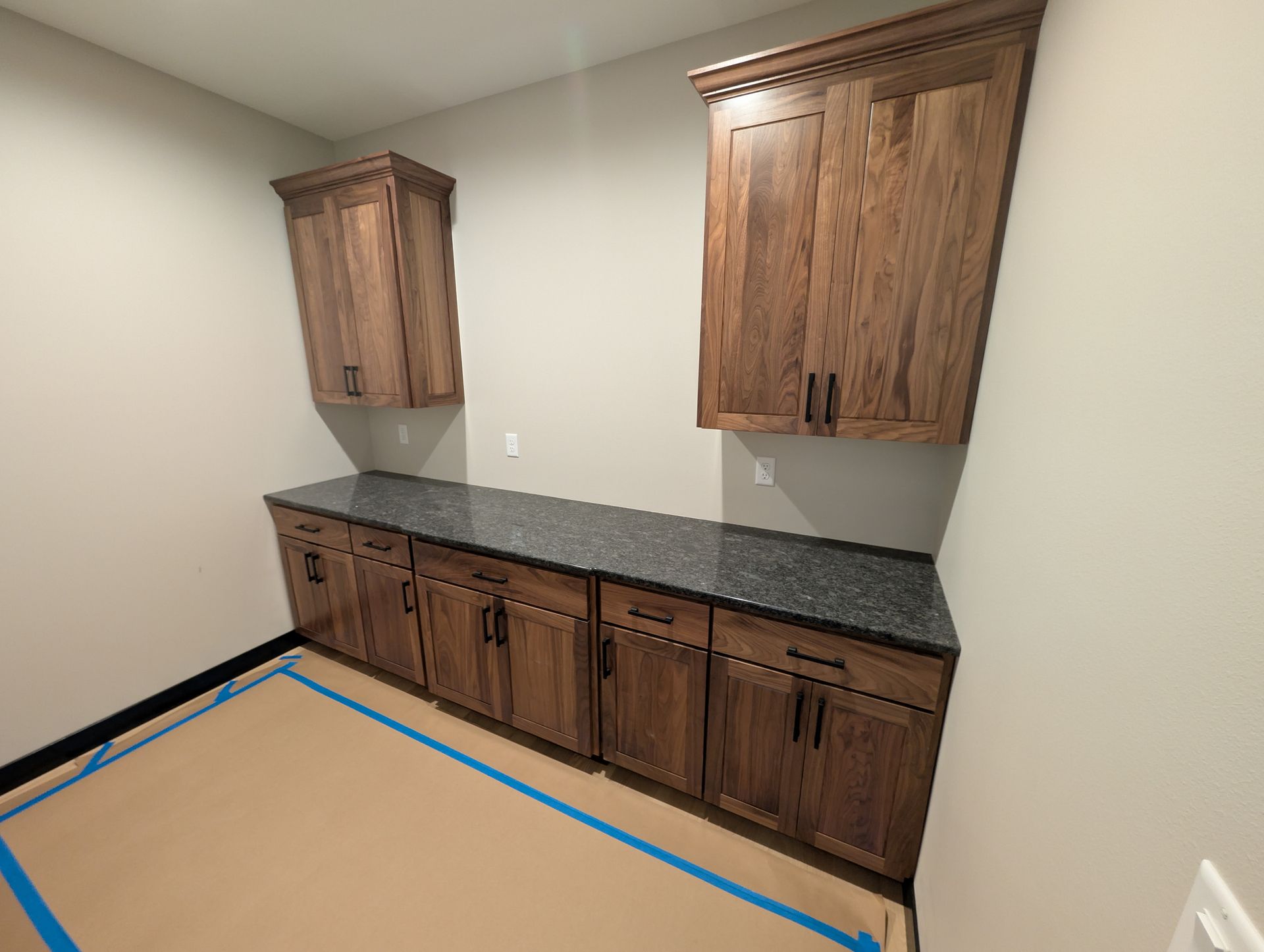 A laundry or kitchen space featuring wood cabinets, dark granite countertops, and light walls with floor protection tape.