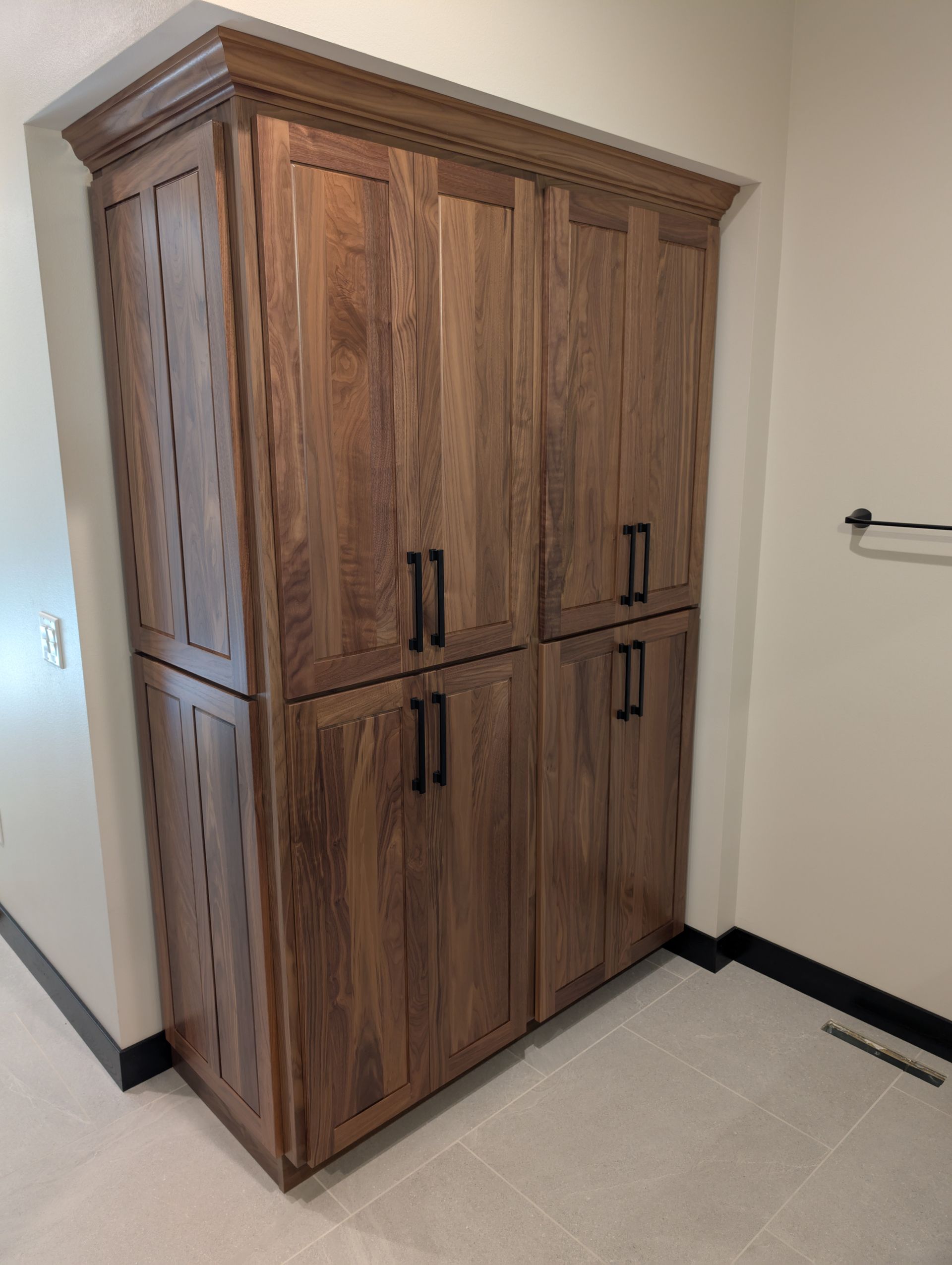A tall, wooden cabinet with four cupboard doors and dark handles, built into an alcove in a room with light tile flooring.
