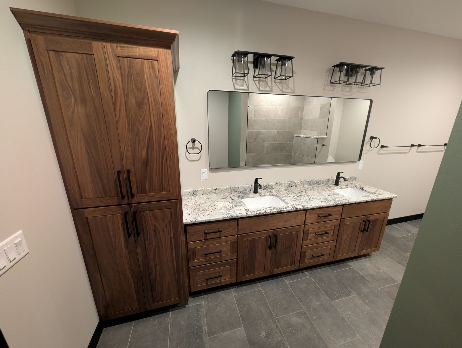 A modern bathroom featuring a double vanity, granite countertop, tall dark wood cabinet, large mirror, and gray floor tiles.