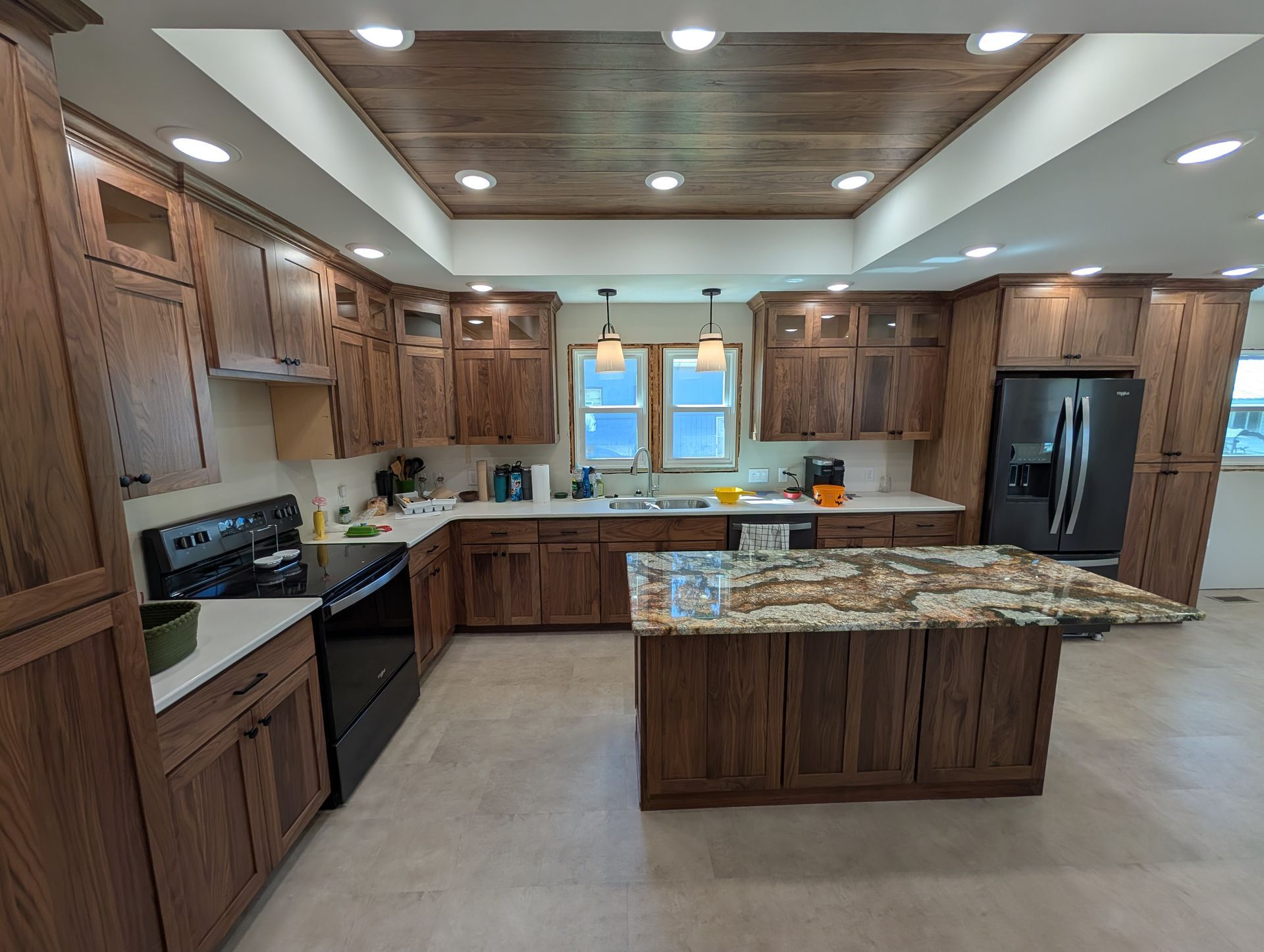 A modern kitchen featuring rich walnut cabinets, a central granite-topped island, dark appliances, and a recessed ceiling.