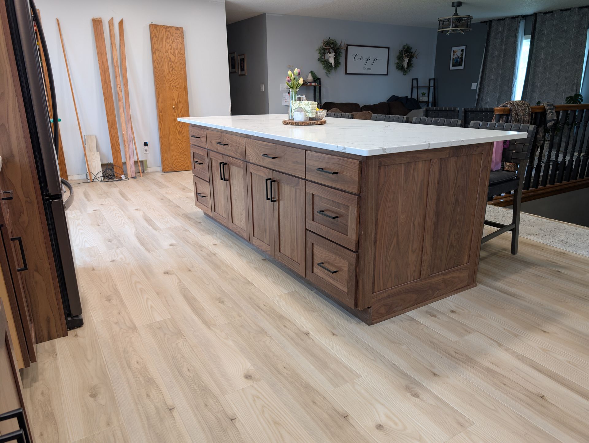 A wooden kitchen island with a white countertop stands in a room with light-colored flooring and an open-plan living area.