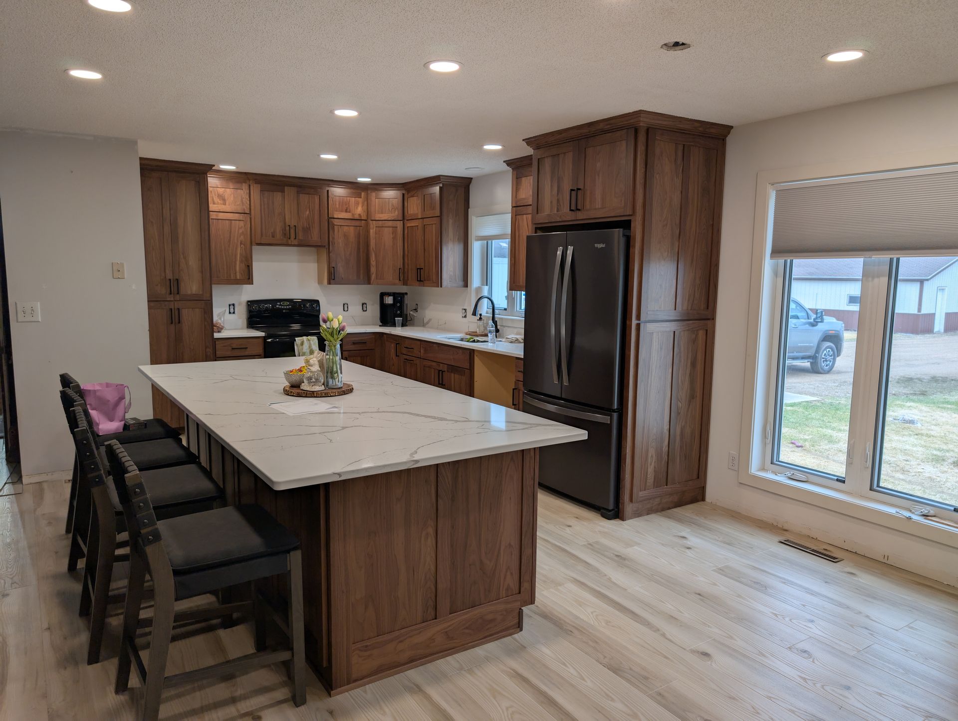 A modern kitchen with wood cabinets, a white marble island with three bar stools, and a black refrigerator.