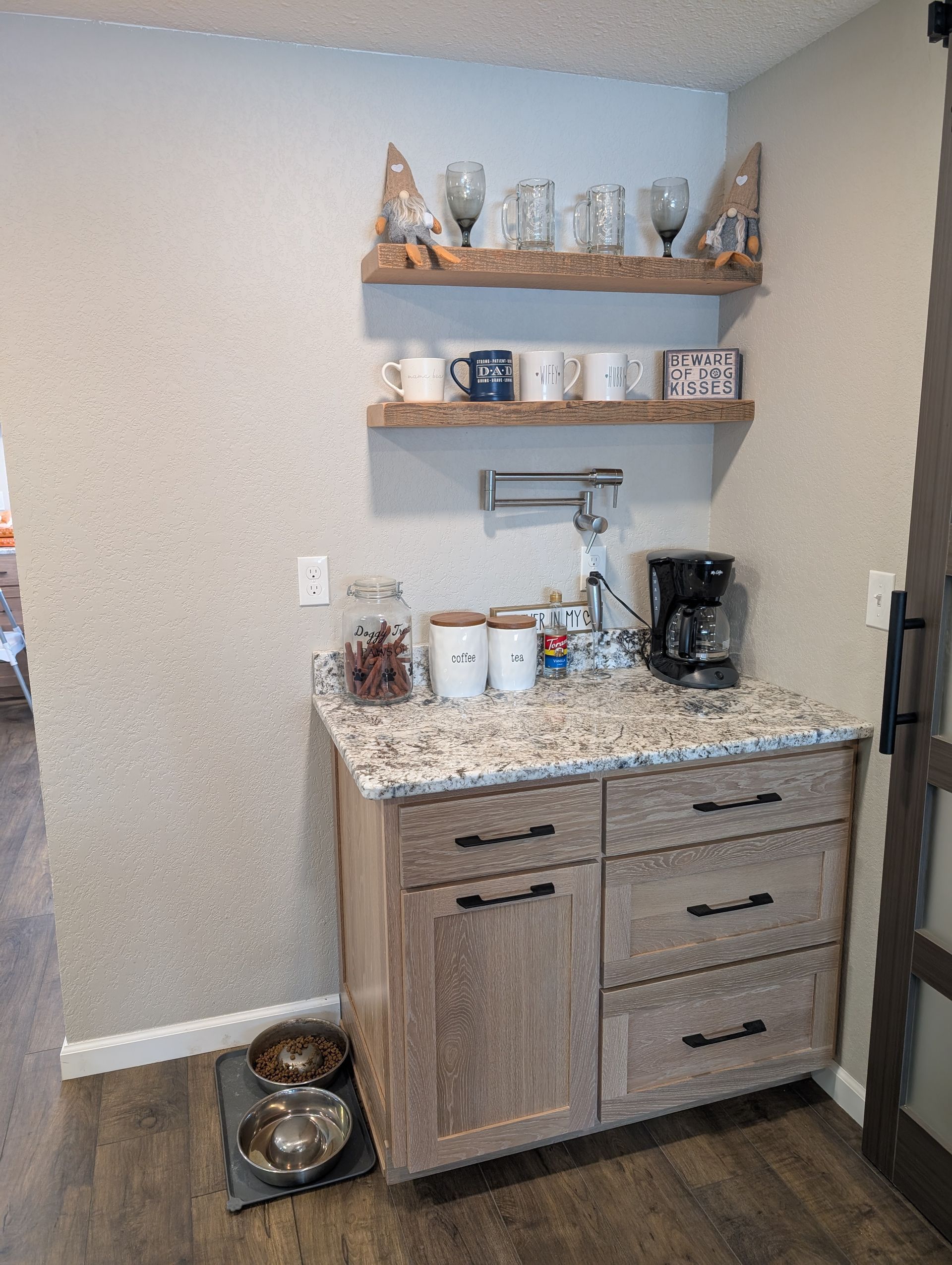 A kitchen coffee station with wood cabinets, stone countertops, two floating shelves, a coffee maker, and pet bowls.