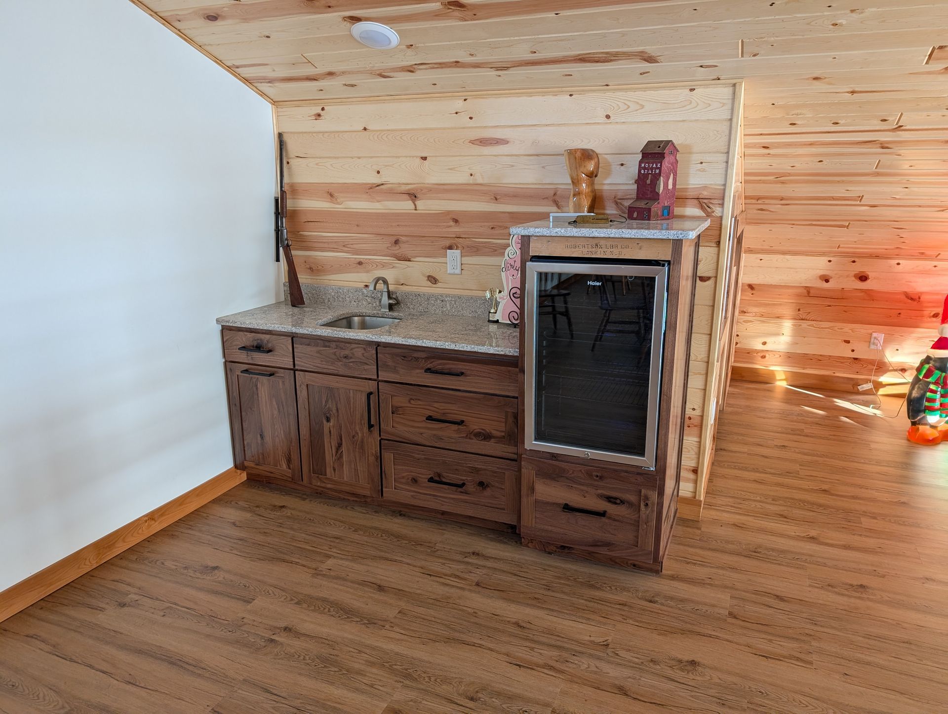 A wet bar with rustic wood cabinets and granite countertops sits against a wood-paneled wall in a room with wood flooring.