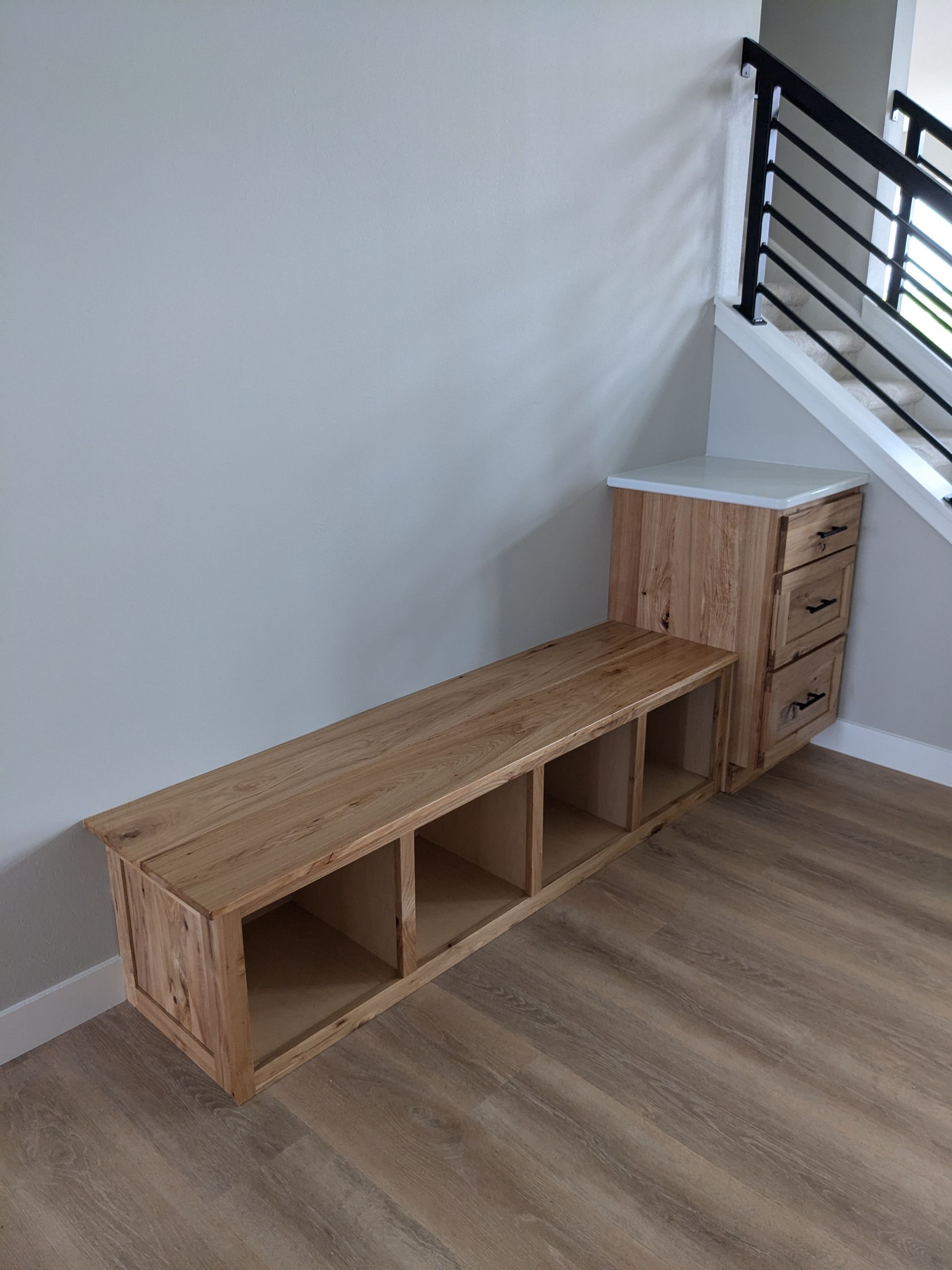 Custom wooden storage bench with open cubbies and a three-drawer cabinet against a light grey wall near a staircase.