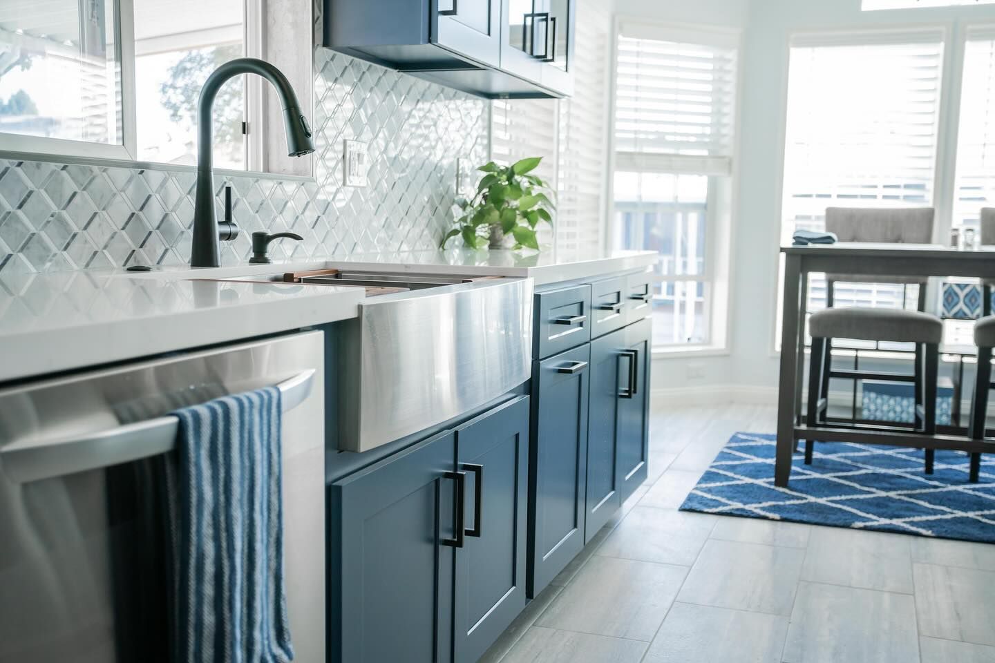 Blue kitchen with stainless steel sink, cabinets, and backsplash. Dining table, rug, and window.