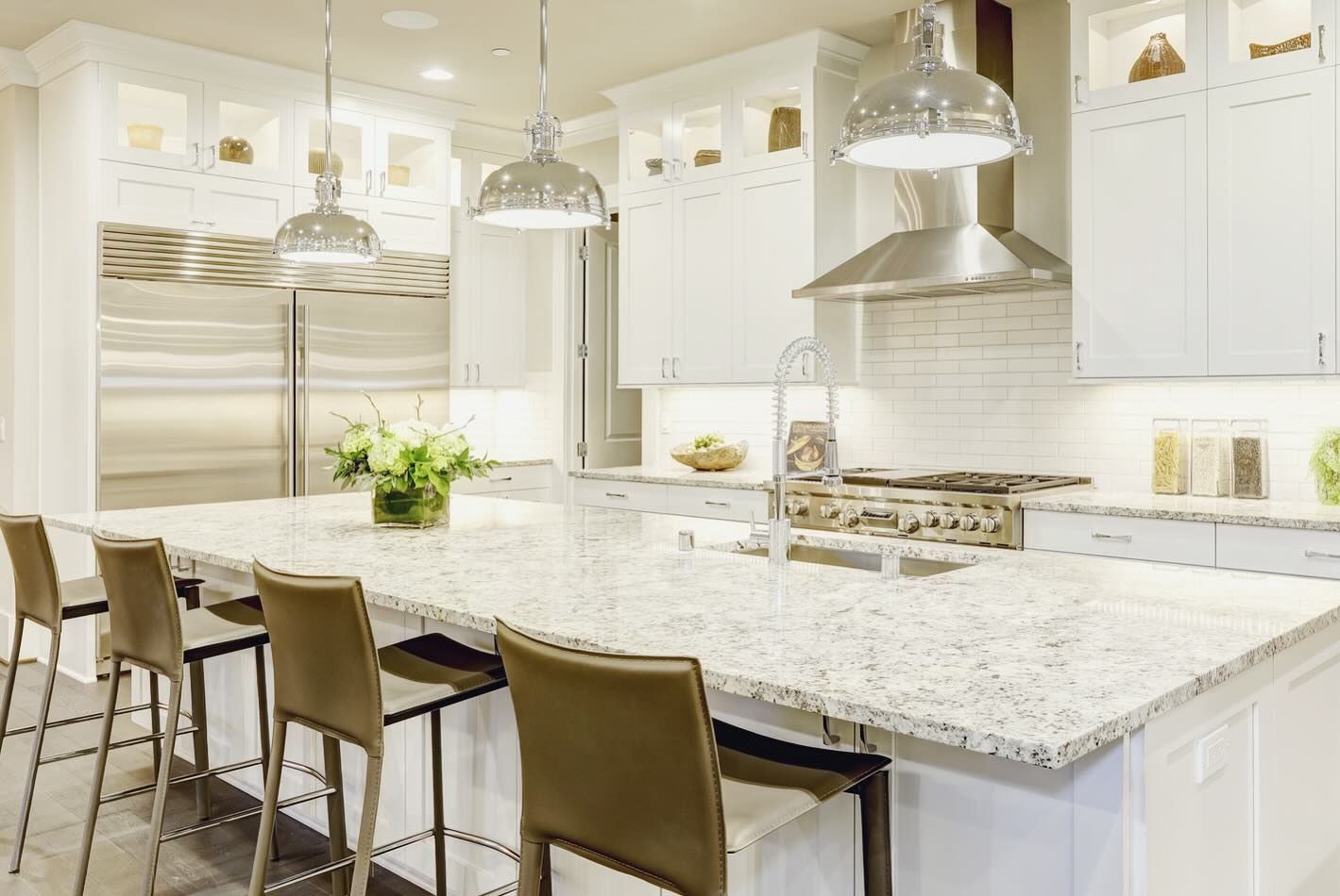 A bright white kitchen with a large island, granite countertops, and metal pendant lights.