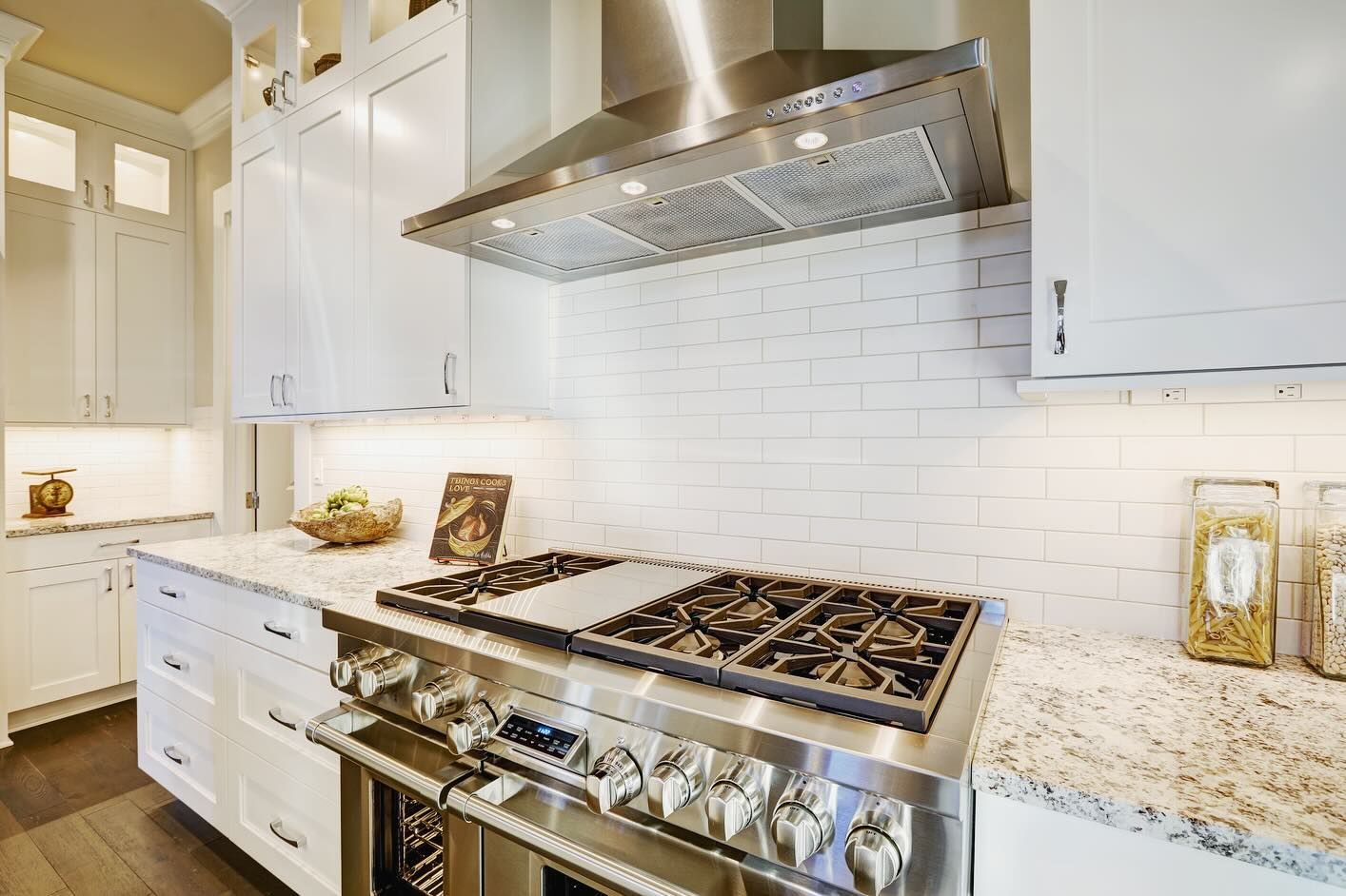 Stainless steel gas range in a bright white kitchen with granite countertops and white brick backsplash.