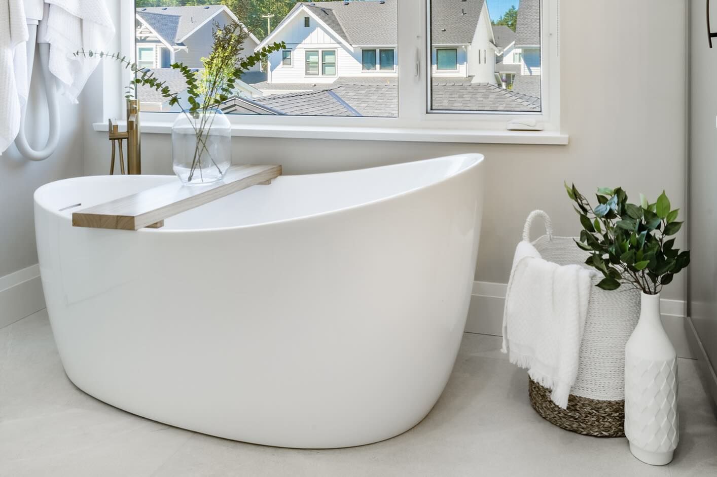 White modern soaking tub in a bathroom with a window, greenery, and a basket with towels.