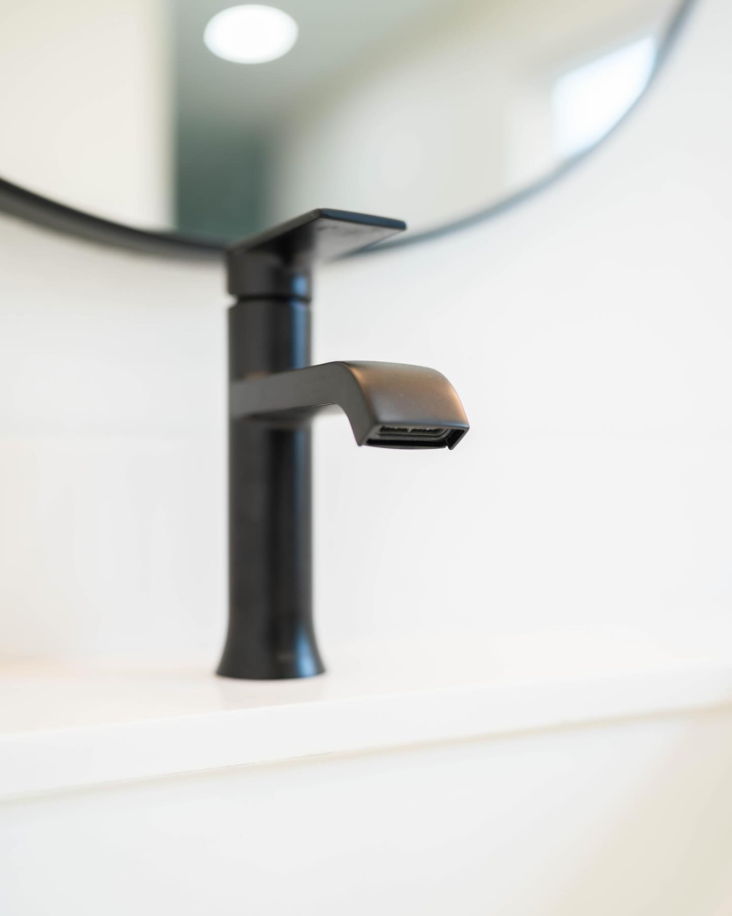 Black faucet in a white bathroom, with a round mirror in the background.