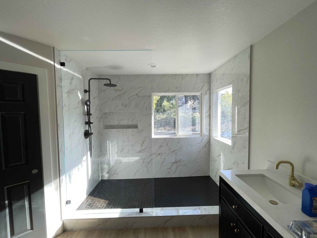 Bathroom remodel: marble-tiled shower, black fixtures, glass door, vanity, and black door on left.