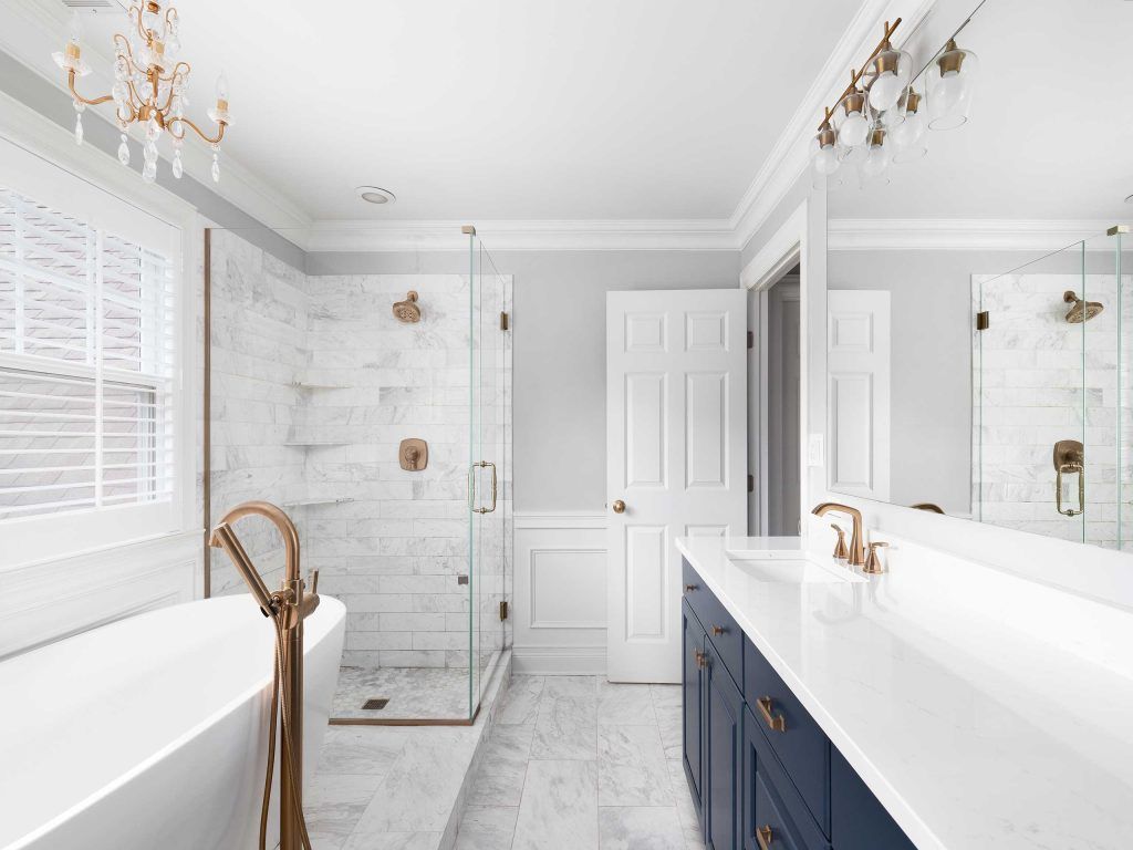A modern bathroom with marble tile, glass shower, blue vanity, white tub, and gold fixtures.