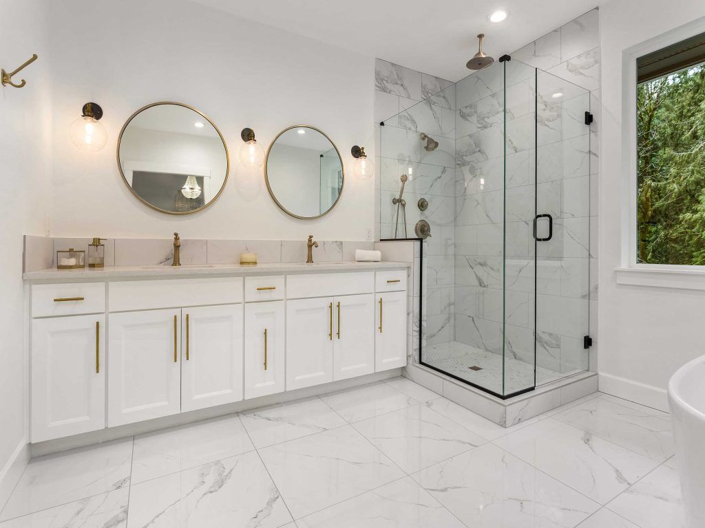 Bright, modern bathroom with white marble and gold accents. Features a double vanity, large shower, and large window.