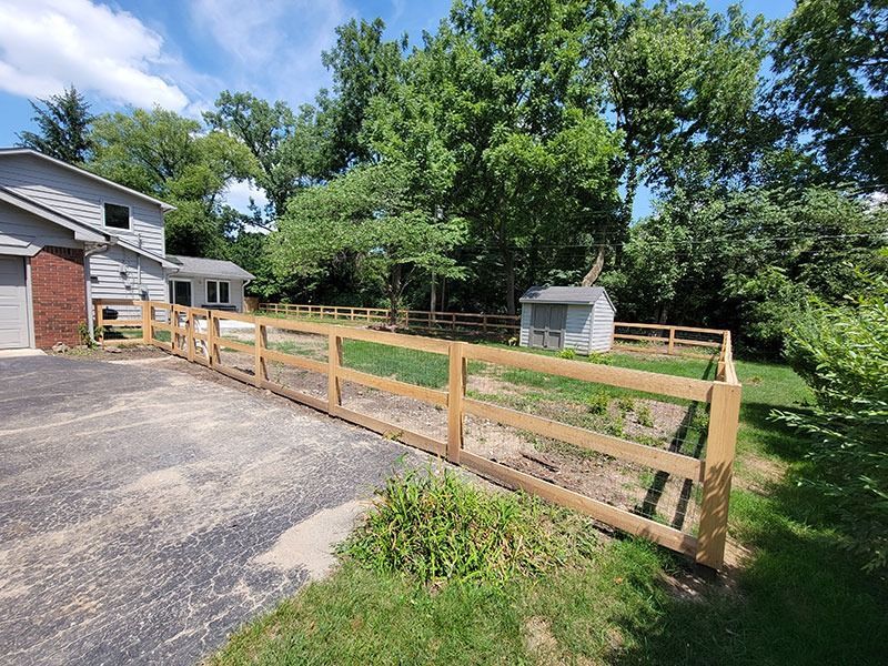 Wooden fence surrounds a yard with a shed, trees, and a house on a sunny day.