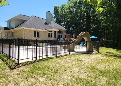A backyard with a pool, fence, slide, and house with a dark roof. Green grass and trees in the background.