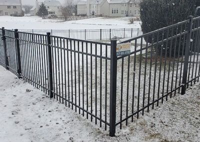Black metal fence surrounding a yard in the snow. Houses in the background.