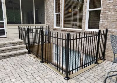 Black fenced-in sunken area on a brick patio. Glass window in the sunken area. Brown brick wall and a screened-in porch are in the background.
