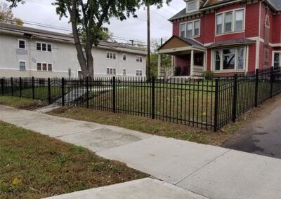 Black metal fence borders a sidewalk and grass. Red brick house in background.