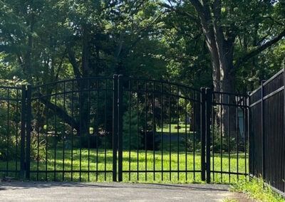 Black wrought-iron gate in a driveway, leading to a grassy yard surrounded by trees.