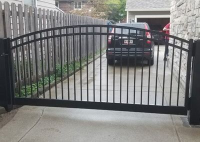 Black metal driveway gate, arched top, closed. Car parked behind gate. Concrete driveway.