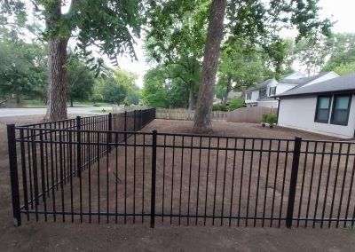 Black metal fence enclosing a small yard between two trees and a house.