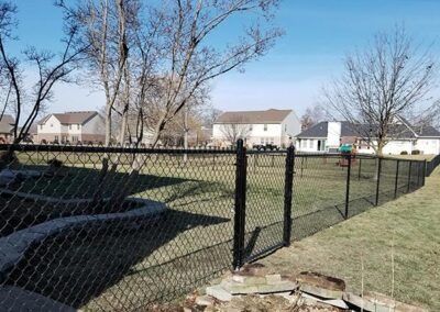 Black chain-link fence encloses a backyard with a gate, trees, and houses in the background on a sunny day.
