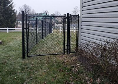 Black chain link fence and gate alongside a house, on a grassy lawn.