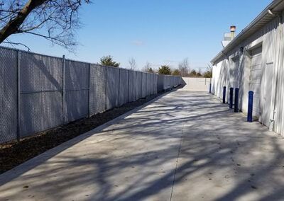 Chain-link fence borders a concrete path beside a white building with blue bollards on a sunny day.