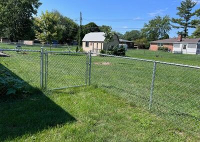 Chain-link fence with gate in grassy backyard, shed and houses in background, under a blue sky.