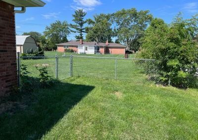 Grassy backyard with chain-link fence, brick building on the left, other houses in the background under a blue sky.